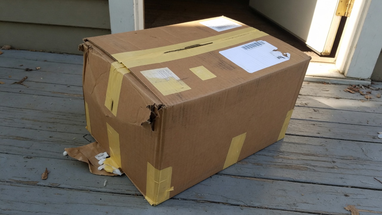 門廊木地板上一個邊角擠壓、膠帶還在的牛皮紙箱,空白面單